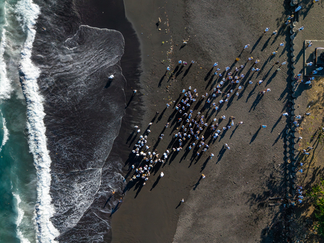 Aerial view of Ngelarung Sea Offering Ceremony at Pura Segara Goa Lawah with people in traditional white clothing gathering on the black sand beach in Dawan Bali Indonesia.