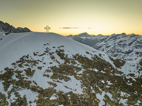 Aerial view of the Vallespluga mountain peak featuring a metal summit cross surrounded by snow covered rocky slopes and distant alpine peaks under a golden sunset sky in Lombardia Italy. Aerial view of the Vallespluga mountain peak featuring a metal summit cross surrounded by snow covered rocky slopes and distant alpine peaks under a golden sunset sky in Lombardia Italy.