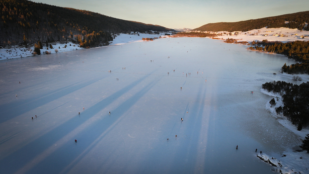 Aerial view of a frozen lake reflecting the golden hour s glow as people skate across its vast icy expanse framed by snow dusted forests Les Rousses Bourgogne Franche Comté France. Aerial view of a frozen lake reflecting the golden hour s glow as people skate across its vast icy expanse framed by snow dusted forests Les Rousses Bourgogne Franche Comté France.