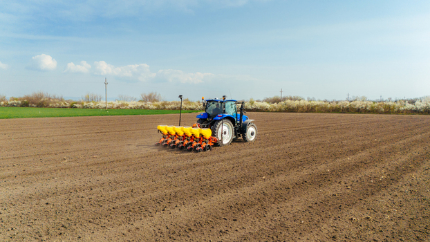 Aerial view of a blue tractor with a yellow seeder trailing behind it working on a vast ploughed field under a bright sky Sremska Mitrovica Vojvodina Serbia. Aerial view of a blue tractor with a yellow seeder trailing behind it working on a vast ploughed field under a bright sky Sremska Mitrovica Vojvodina Serbia.