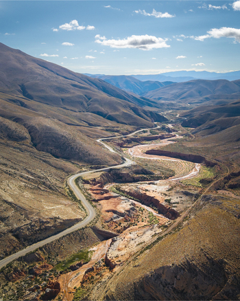 Aerial view of a winding asphalt road snaking through a deep arid mountain valley with rocky slopes sparse vegetation and a dry riverbed in Argentina. Aerial view of a winding asphalt road snaking through a deep arid mountain valley with rocky slopes sparse vegetation and a dry riverbed in Argentina.
