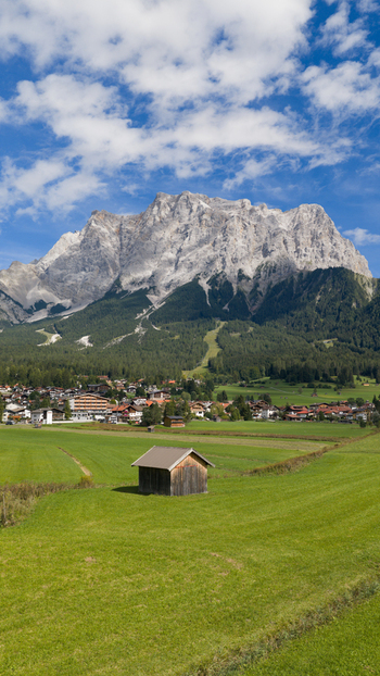 Aerial view of a small wooden hut in a lush green meadow with the village of Grainau and the towering Zugspitze mountain in the background in Grainau Bayern Germany. Aerial view of a small wooden hut in a lush green meadow with the village of Grainau and the towering Zugspitze mountain in the background in Grainau Bayern Germany.