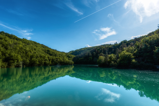 View of the turquoise waters of a tranquil lake surrounded by lush green hills and dense forest under a clear blue sky in Pievebovigliana Marche Italy. View of the turquoise waters of a tranquil lake surrounded by lush green hills and dense forest under a clear blue sky in Pievebovigliana Marche Italy.