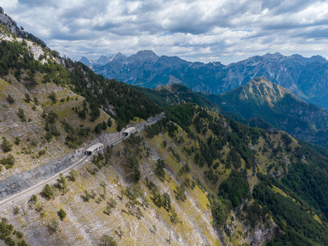 Aerial view of the winding mountain road with tunnels carved into the rocky slopes of the Albanian Alps surrounded by forests and dramatic peaks in Shkoder Shkoder County Albania. Aerial view of the winding mountain road with tunnels carved into the rocky slopes of the Albanian Alps surrounded by forests and dramatic peaks in Shkoder Shkoder County Albania.