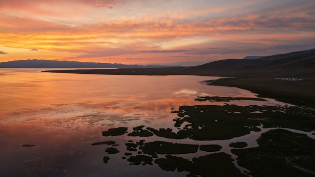 Aerial view of Song Kul lake at sunset with vibrant orange and pink clouds reflecting on the calm water surrounded by rolling hills and remote grasslands in Naryn Region Kyrgyzstan. Aerial view of Song Kul lake at sunset with vibrant orange and pink clouds reflecting on the calm water surrounded by rolling hills and remote grasslands in Naryn Region Kyrgyzstan.