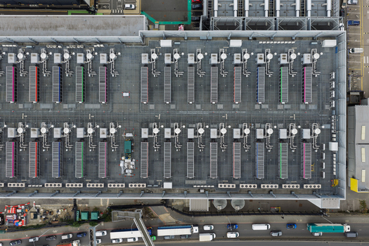 Aerial view of a large data center roof featuring rows of cooling units and ventilation systems with a busy road and vehicles visible at the bottom edge United Kingdom.