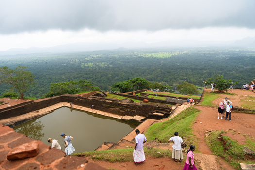 Sri Lanka 21 February 2026 View of the ancient Sigiriya rock fortress ruins featuring a large water tank and tourists overlooking the vast green jungle landscape.