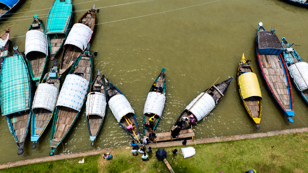 Aerial view of Baish Mowza Sarak Bazar with wooden boats moored along the riverbank people gathering near the water and green grass in Nabinagar Chittagong Division Bangladesh. Aerial view of Baish Mowza Sarak Bazar with wooden boats moored along the riverbank people gathering near the water and green grass in Nabinagar Chittagong Division Bangladesh.