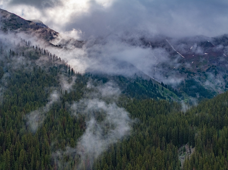 View of misty mountain peaks rising above a dense evergreen forest with low hanging clouds rolling over the slopes under a moody sky Colorado United States.