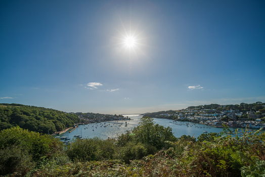 View of the Fowey River estuary with boats moored in the water and the historic town of Fowey nestled on the hillside under a bright sun in Fowey Cornwall United Kingdom. View of the Fowey River estuary with boats moored in the water and the historic town of Fowey nestled on the hillside under a bright sun in Fowey Cornwall United Kingdom.