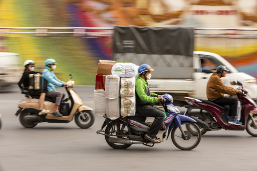 Hanoi Vietnam 11 November 2025 View of motorcyclists navigating busy city streets with heavy cargo loads and wearing face masks captured with motion blur in the background.