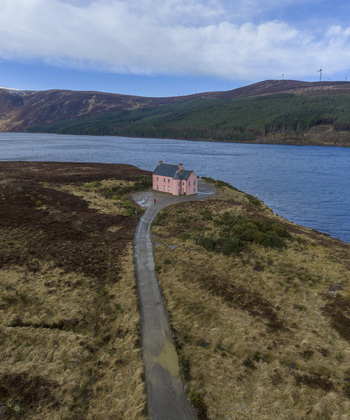 Aerial view of The Pink House isolated on a peninsula by a lake surrounded by rugged moorland and hills with wind turbines in Dingwall Scotland United Kingdom. Aerial view of The Pink House isolated on a peninsula by a lake surrounded by rugged moorland and hills with wind turbines in Dingwall Scotland United Kingdom.