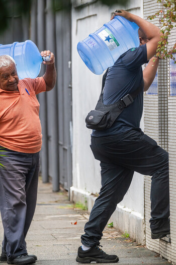 Rio de Janeiro Brazil 20 May 2026 View of two men carrying large blue plastic water jugs on their shoulders while walking along a sidewalk next to a white wall. Rio de Janeiro Brazil 20 May 2026 View of two men carrying large blue plastic water jugs on their shoulders while walking along a sidewalk next to a white wall.