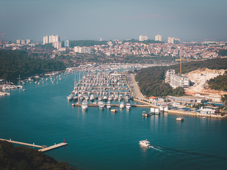 Aerial view of azure waters cradling a dense harbor filled with gleaming white yachts contrasting with the verdant hills and distant city skyline Pula Istria County Croatia.