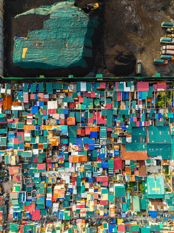 Aerial view of the dense Happyland slum in Tondo featuring colorful rooftops adjacent to an industrial site with a large green tarp and dark soil piles in Manila Metro Manila Philippines.