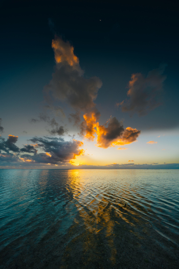 View of Aitutaki lagoon at sunset with golden clouds reflecting on the calm water in Cook Islands. View of Aitutaki lagoon at sunset with golden clouds reflecting on the calm water in Cook Islands.