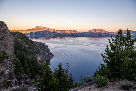 View of a serene blue lake reflecting the soft glow of the sunrise surrounded by towering cliffs and lush green forests Crater Lake Oregon United States. View of a serene blue lake reflecting the soft glow of the sunrise surrounded by towering cliffs and lush green forests Crater Lake Oregon United States.