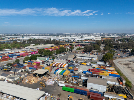Aerial view of a busy shipping container yard with colorful cargo containers industrial buildings and heavy machinery under a clear blue sky in Carson California United States.