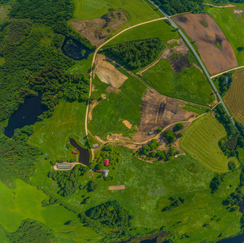 Aerial view of a rural landscape featuring lush green fields patches of forest small ponds and a dirt road winding past farm buildings in Latvia. Aerial view of a rural landscape featuring lush green fields patches of forest small ponds and a dirt road winding past farm buildings in Latvia.