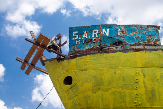 Keraniganj Bangladesh 04 September 2024 View of a worker sitting on a wooden scaffold while repairing the bow of a large rusty yellow ship at Shipyard Dhaka under a blue sky.