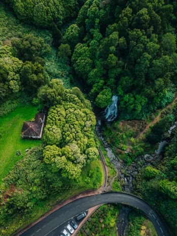 Aerial view of a winding road curves near a verdant forest adjacent to a quaint building and a cascading waterfall Achada Açores Portugal. Aerial view of a winding road curves near a verdant forest adjacent to a quaint building and a cascading waterfall Achada Açores Portugal.