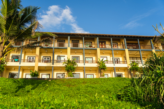 View of a modern three story hotel building with yellow walls and wooden balconies surrounded by lush palm trees and green grass under a blue sky in Galle Southern Province Sri Lanka.