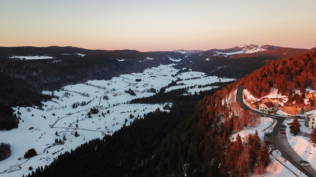 Aerial view of Col de la Faucille featuring a snowy mountain valley with pine forests and a winding road illuminated by sunset light in Gex Auvergne Rhone Alpes France.