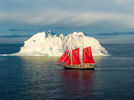 Aerial view of a sailboat with vibrant red sails sailing past a massive iceberg in the Arctic waters of Greenland. Aerial view of a sailboat with vibrant red sails sailing past a massive iceberg in the Arctic waters of Greenland.