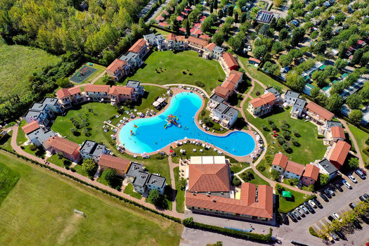 Aerial view of a resort complex with people swimming in a large blue pool surrounded by colorful houses and green lawns near Lake Garda Italy. Aerial view of a resort complex with people swimming in a large blue pool surrounded by colorful houses and green lawns near Lake Garda Italy.
