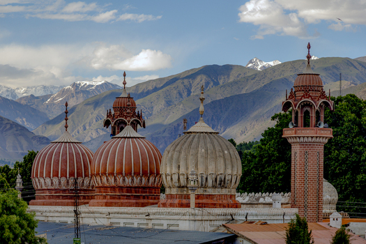 View of the historic Shahi Mosque with its distinctive red and white domes and minaret set against the snow capped Hindu Kush mountains in Chitral Khyber Pakhtunkhwa Pakistan.