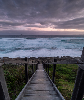 View of the wooden staircase leading down to the rocky coastline during a cloudy sunset with waves crashing against the shore at Sandpatch Western Australia Australia. View of the wooden staircase leading down to the rocky coastline during a cloudy sunset with waves crashing against the shore at Sandpatch Western Australia Australia.