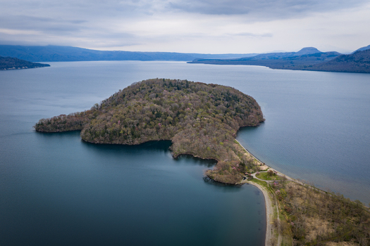 Aerial view of the forested Nakajima Island in the middle of the volcanic Lake Kussharo with calm blue waters and a cloudy sky in Teshikaga Hokkaido Japan. Aerial view of the forested Nakajima Island in the middle of the volcanic Lake Kussharo with calm blue waters and a cloudy sky in Teshikaga Hokkaido Japan.