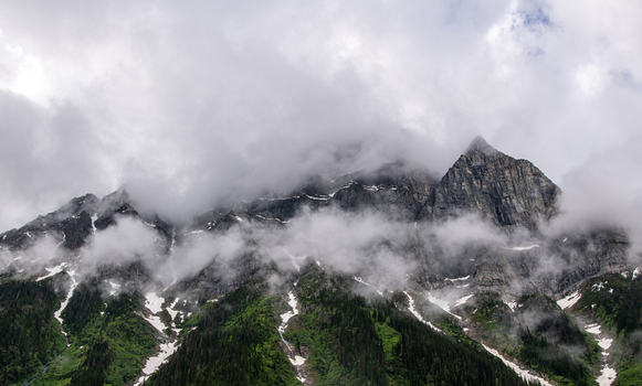 View of jagged peaks rise majestically shrouded in ethereal mist patches of snow clinging to steep slopes green vegetation contrasting with the grey rock Revelstoke Columbia Shuswap Canada.