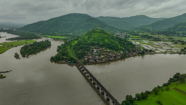 Aerial view of a railway bridge crossing a river near a village nestled on a lush green hill surrounded by agricultural fields and rolling mountains Mahad Maharashtra India.