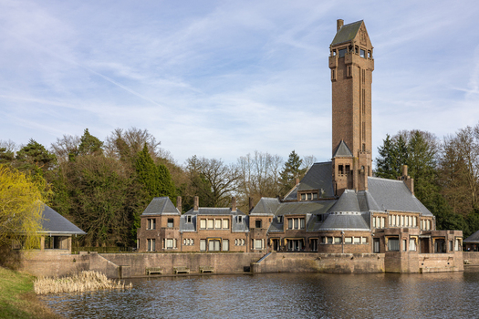 View of Country Residence Museum Jachthuis Sint Hubertus a historic brick hunting lodge with a tall tower overlooking a calm pond in Hoenderloo Gelderland Netherlands.