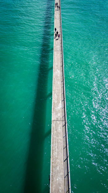Aerial view of a long pier stretching into the turquoise sea casting a dramatic shadow as people stroll along its length Roscoff Brittany France. Aerial view of a long pier stretching into the turquoise sea casting a dramatic shadow as people stroll along its length Roscoff Brittany France.