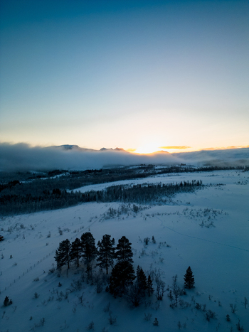 Aerial view of the snowy winter landscape on Kvaloya island with pine trees in the foreground low hanging clouds and a golden sunrise over the mountains in Troms Norway.