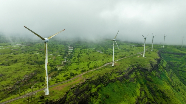 Aerial view of wind turbines standing on a lush green ridge shrouded in mist with rolling hills and a winding road in the background Igatpuri Maharashtra India. Aerial view of wind turbines standing on a lush green ridge shrouded in mist with rolling hills and a winding road in the background Igatpuri Maharashtra India.