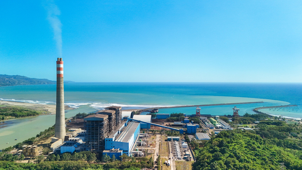 Aerial view of a power plant with a tall chimney emitting smoke situated along the coastline with blue sea and lush green hills in Indonesia.