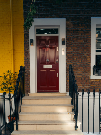 London United Kingdom 21 June 2025 View of a vibrant red door framed by stark white trim steps leading up the dark railings contrasting against the aged brick and yellow wall. London United Kingdom 21 June 2025 View of a vibrant red door framed by stark white trim steps leading up the dark railings contrasting against the aged brick and yellow wall.