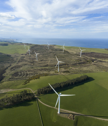 Aerial view of a windfarm with white turbines standing on green fields and moorland near the coast under a blue sky Scotland United Kingdom. Aerial view of a windfarm with white turbines standing on green fields and moorland near the coast under a blue sky Scotland United Kingdom.