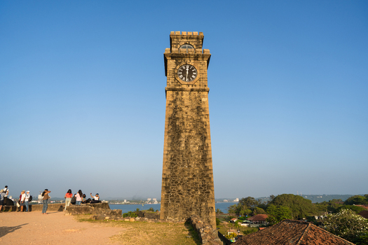 View of the historic Galle Fort Clock Tower with tourists standing on the ramparts under a clear blue sky in Galle Southern Province Sri Lanka.