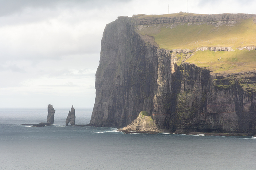 View of the dramatic sea cliffs and the Risin og Kellingin sea stacks rising from the ocean under a cloudy sky in the Faroe Islands.
