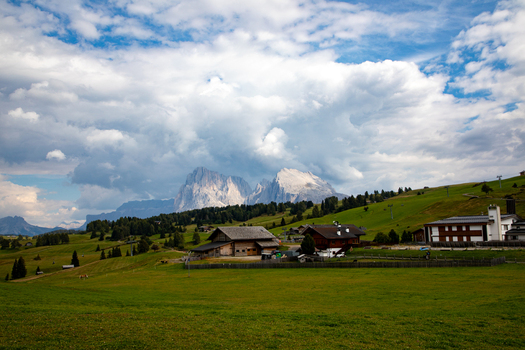 View of traditional wooden chalets in a lush green alpine meadow with the jagged peaks of the Dolomites under a cloudy sky in Rocca Pietore Veneto Italy. View of traditional wooden chalets in a lush green alpine meadow with the jagged peaks of the Dolomites under a cloudy sky in Rocca Pietore Veneto Italy.