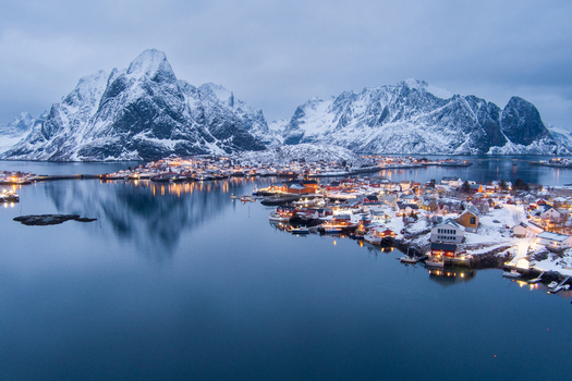 Aerial view of the snowy fishing village of Reine in Lofoten with traditional red cabins snow covered mountains and calm water reflecting evening lights in Nordland Norway.