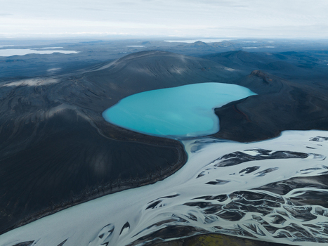 Aerial view of the vibrant turquoise lake embraced by the stark black volcanic landscape with glacial rivers carving intricate patterns below Highlands Iceland.