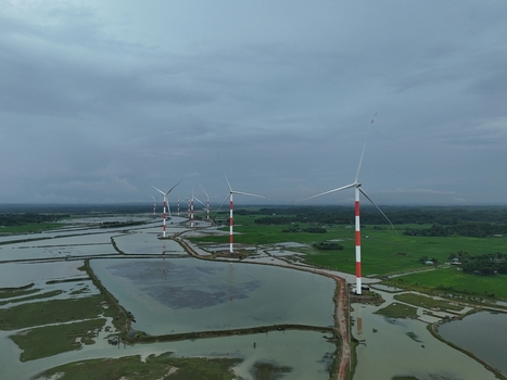 Aerial view of the Khurushkul wind power plant with red and white turbines standing in flooded wetlands and green fields under a cloudy sky in Cox s Bazar Chittagong Division Bangladesh. Aerial view of the Khurushkul wind power plant with red and white turbines standing in flooded wetlands and green fields under a cloudy sky in Cox s Bazar Chittagong Division Bangladesh.