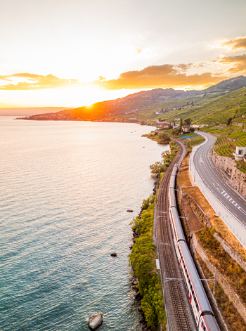 Aerial view of a passenger train traveling along the scenic shoreline of Lake Geneva passing through the terraced vineyards of Lavaux at sunset in Bourg en Lavaux Vaud Switzerland. Aerial view of a passenger train traveling along the scenic shoreline of Lake Geneva passing through the terraced vineyards of Lavaux at sunset in Bourg en Lavaux Vaud Switzerland.