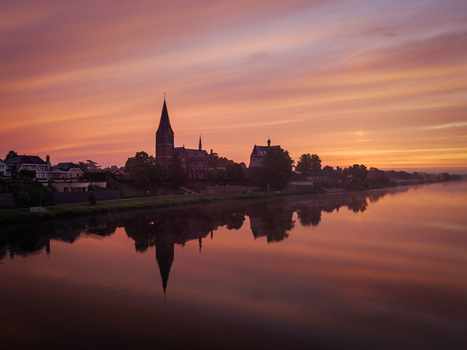 View of the silhouette of the historic church and village buildings reflected in the calm river Maas during a vibrant sunset in Kessel Limburg Netherlands. View of the silhouette of the historic church and village buildings reflected in the calm river Maas during a vibrant sunset in Kessel Limburg Netherlands.