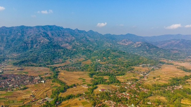 Aerial view of Ngadiharjo village surrounded by terraced rice fields lush green hills and rural houses under a clear blue sky in Borobudur Central Java Indonesia.